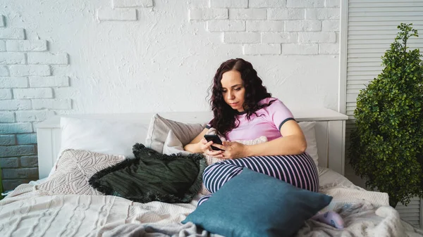 Young woman learning remotely by smartphone while lying on pillows in bed.
