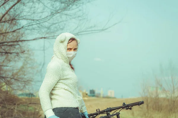Young woman in medical mask and gloves stands with bicycle, holding on to rudder in countryside. Female protecting yourself from diseases on walk. Concept of threat of coronavirus epidemic infection.
