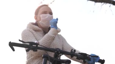 Tıbbi maskeli ve eldivenli genç bir kadın öksürüyor, kırsalda bisiklet dümenine tutunuyor. Yürüyüş sırasında kendini hastalıklardan koruyan bir kadın. Koronavirüs salgını tehdidi kavramı.