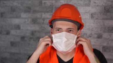 Tulumlu erkek inşaat işçisi, gri tuğla duvarın arka planındaki tıbbi maskeyi çıkarıyor.