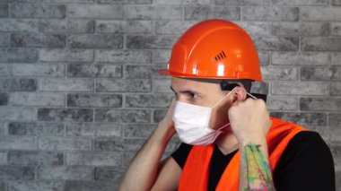Tulumlu erkek inşaat işçisi, gri tuğla duvarın arka planındaki tıbbi maskeyi çıkarıyor.