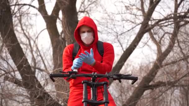 Joven con máscara médica poniéndose guantes, sentado en bicicleta en el campo. Hombre protegiéndose de las enfermedades a pie. Concepto de amenaza de infección epidémica por coronavirus . — Vídeo de stock