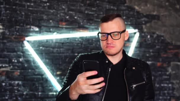 Un tânăr cu ochelari răsucind prin știri în telefonul mobil pe peretele de cărămidă de fundal cu lămpi strălucitoare. Învățarea brutală a bărbaților de la distanță prin smartphone în timp ce stați împotriva peretelui iluminat . — Videoclip de stoc