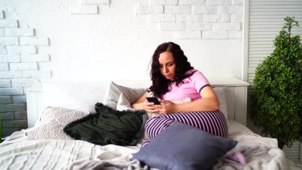 Взрослая очаровательная женщина в пижаме листает различные новости в мобильном телефоне в спальне. Молодая женщина дистанционно учится на смартфоне, лежа на подушках в постели. — стоковое видео