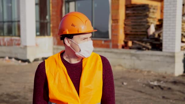 Portret van een mannelijke bouwvakker met medisch masker en overall op de achtergrond van een huis in aanbouw. — Stockvideo