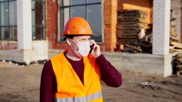 Male construction worker in overalls and medical mask talking about coronavirus on mobile phone on background of house under construction. Concept of threat of infection. — Stock Video