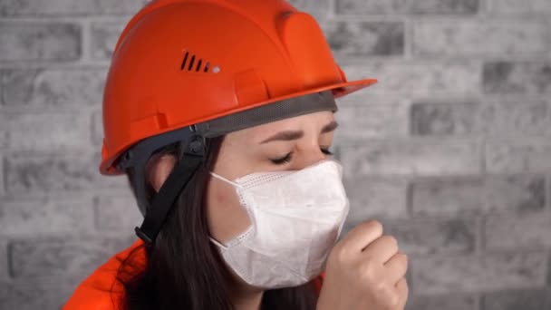 Γυναίκα οικοδόμος με φόρμες και ιατρική μάσκα βήχας στο φόντο του γκρίζου τοίχου από τούβλα. Έννοια της απειλής μόλυνσης. — Αρχείο Βίντεο