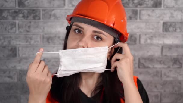 Female construction worker in overalls putting on medical mask on face on background of gray brick wall — Stock Video