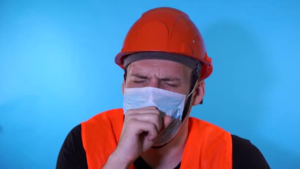 Önlüklü ve maske takmış mavi arka planda öksüren bir inşaat işçisi. Enfeksiyon tehdidi kavramı. — Stok video