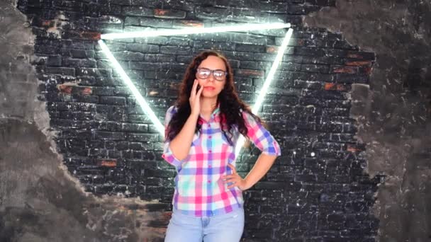 Young woman with glasses in colored plaid shirt posing on background shabby brick wall with glowing lamps. Portrait of charming female holding eyeglasses with her hand against illuminated wall. — Stock Video