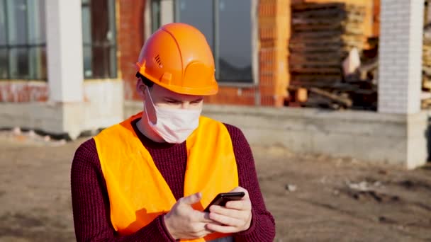 Male construction worker in overalls and medical mask flipping through news about coronavirus in mobile phone on background of house under construction. Concept of threat of infection. — Stock Video