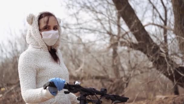 Mujer joven en máscara médica y guantes se para con la bicicleta, aferrándose al timón en el campo. Mujer protegiéndose de las enfermedades a pie. Concepto de amenaza de infección epidémica por coronavirus . — Vídeos de Stock