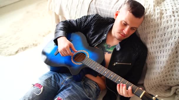 A young man with a tattoo on his chest plays the guitar in the bedroom. Hipster learn to play the guitar. Close-up of a mans hand playing a guitar string. Concept: rock musician — Stock Video