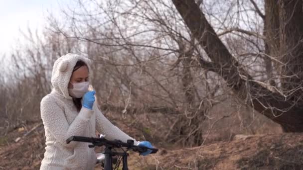 Jeune femme en masque médical et gants toussant, tenant au gouvernail de bicyclette dans la campagne. Femme se protégeant contre les maladies à pied. Concept de menace d'infection par le coronavirus . — Video