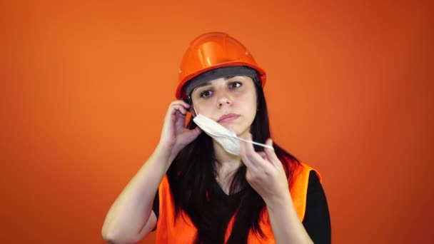 Mujer trabajadora de la construcción en overoles que se pone máscara médica en la cara sobre fondo naranja . — Vídeo de stock