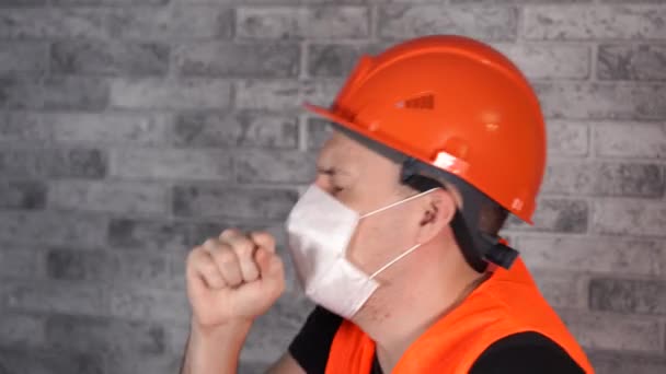 회색 벽돌 벽 앞에서 작업복을 입고 의료용 마스크를 쓴 남성 건설 근로자가 기침을 하고 있습니다. 감염의 위협에 대한 개념. — 비디오