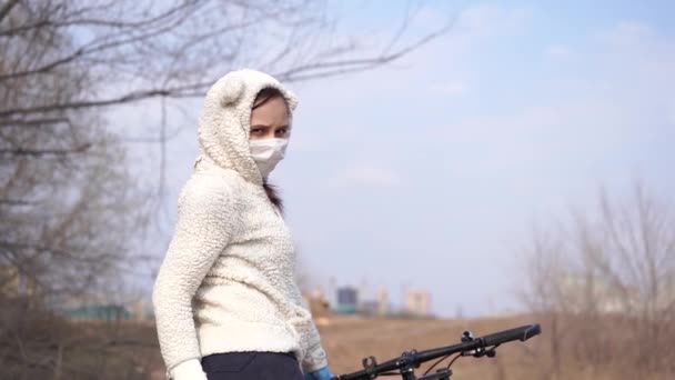 Young woman in medical mask and gloves stands with bicycle, holding on to rudder in countryside. Female protecting yourself from diseases on walk. Concept of threat of coronavirus epidemic infection. — Stock Video