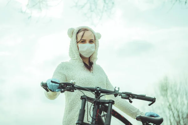 Portrait of young woman in medical mask and gloves with bicycle against sky. Adult female covering face to protect yourself from diseases on walk. Concept of threat of coronavirus epidemic infection.