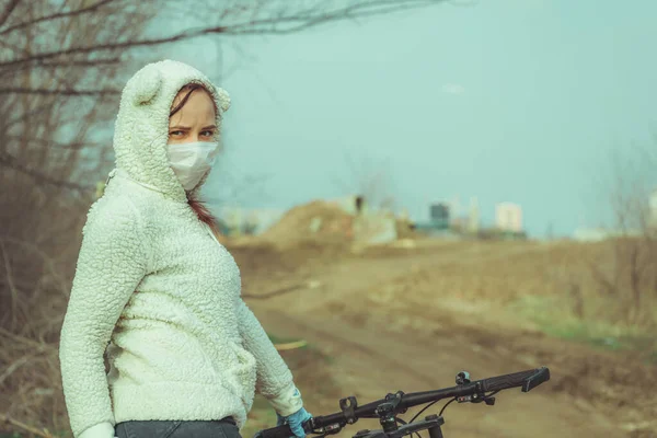 Ung kvinna i medicinsk mask och handskar står med cykel, hålla fast vid rodret på landsbygden. Kvinna som skyddar dig från sjukdomar på promenad. Begreppet hot om infektion med coronavirus. — Stockfoto