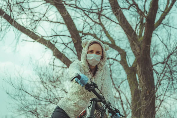 Молодая женщина в медицинской маске и перчатках стоит с велосипедом, держа руль в сельской местности. Женщина защищает себя от болезней во время ходьбы. Концепция угрозы коронавирусной эпидемии . — стоковое фото