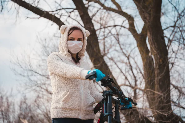 Молода жінка в медичній масці і рукавичках стоїть з велосипедом, тримаючись за кермо в сільській місцевості. Жінка захищає себе від хвороб на прогулянці. Концепція загрози коронавірусної інфекції . — стокове фото