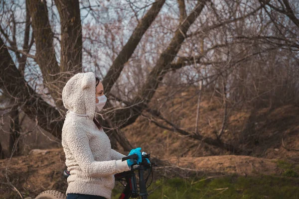 Mladá žena v lékařské masce a rukavicích stojí na kole a drží se kormidla na venkově. Žena, která se chrání před nemocemi na procházce. Koncepce hrozby nákazy koronavirem. — Stock fotografie