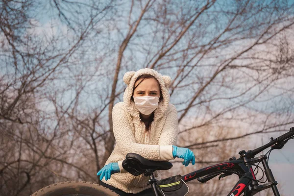 Молода жінка в медичній масці і рукавичках стоїть, спираючись на велосипед в сільській місцевості. Жінка захищає себе від хвороб на прогулянці. Концепція загрози коронавірусної інфекції . — стокове фото