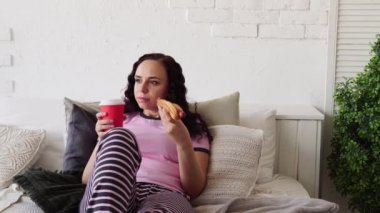 Yatakta sosisli yiyip kahve içen genç bir kadın. Yatakta kahvaltı. Evinize yemek teslimatı. Kadın sıcak bir yatak odasında sulu bir sosisli sandviç yiyor. Konsept: diyet ve fast food günaha teşvik