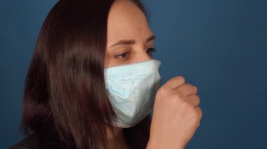 Tıbbi maskeli genç bir kadının portresi öksürüyor, mavi arka planda izole ediliyor. Yetişkin bir kadın kendini hastalıklardan koruyor. Koronavirüs salgını tehdidi kavramı.