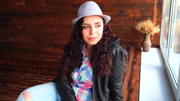 Young beautiful woman in hat and leather jacket sitting by windows. Portrait of pretty female looking out the window in daytime. — Stock Video