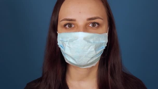 Портрет молодої жінки в медичній масці на її обличчі на синьому фоні. Доросла жінка захищає себе від хвороб. Концепція загрози коронавірусної інфекції . — стокове відео