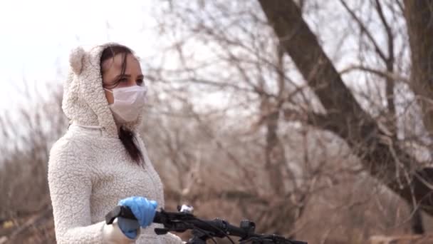 Jonge vrouw met medisch masker en handschoenen staat met de fiets aan het roer op het platteland. Vrouw die zich beschermt tegen ziektes tijdens de wandeling. Concept van dreiging van een besmetting met het coronavirus. — Stockvideo