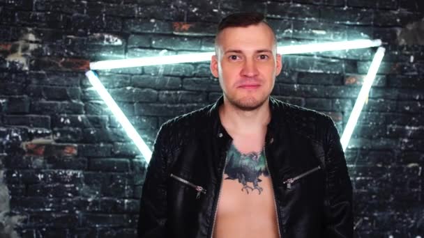 Young man in leather jacket on naked body with tattoo on chest smiling on background brick wall with glowing lamps. Portrait of adult charming male against illuminated wall. — Stock Video