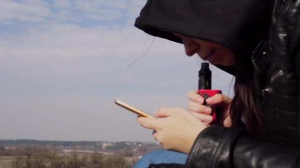 Close-up van jonge vrouw in capuchon dampen en flippen door nieuws in mobiele telefoon, zittend op hoge grond op het platteland. — Stockvideo