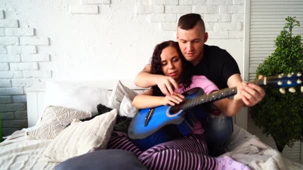 Jeune couple amoureux assis et s'amusant avec un instrument de musique sur le lit dans la chambre. Homme enseignant à la femme à jouer de la guitare à la maison. — Video
