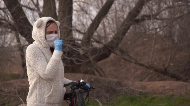 Young woman in medical mask and gloves coughing, holding on to rudder of bicycle in countryside. Female protecting yourself from diseases on walk. Concept of threat of coronavirus epidemic infection. — Stock Video