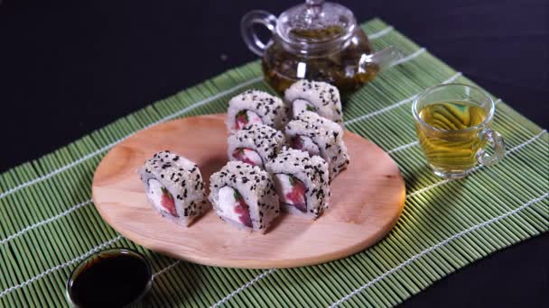 Deliciosos rollos sobre tabla de madera con salsa de soja y té verde sobre servilleta de bambú decorativa. Primer plano de sabrosos rollos de sushi con palillos en la tabla de cortar sobre fondo negro — Vídeo de stock