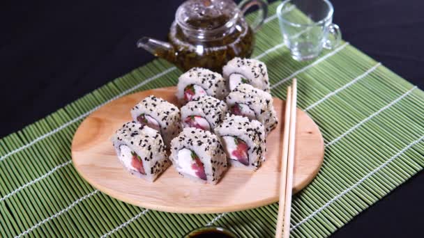 Delicious rolls on wooden board with soy sauce and green tea on decorative bamboo napkin. Close up of tasty sushi rolls with chopsticks on cutting board with steam on black background — Stock Video