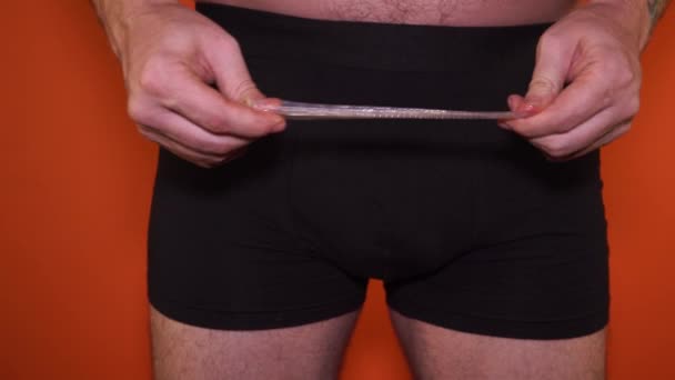 Großaufnahme eines Mannes in Unterwäsche, der ein Kondom auf orangefarbenem Hintergrund hält. Mann in Unterwäsche mit Kondom und Verhütungsmittel. Verhütung - Mann in Unterwäsche — Stockvideo
