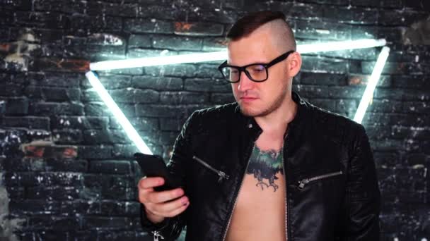 Un tânăr cu ochelari răsucind prin știri în telefonul mobil pe peretele de cărămidă de fundal cu lămpi strălucitoare. Învățarea masculină adultă de la distanță prin smartphone în timp ce stă împotriva peretelui iluminat . — Videoclip de stoc