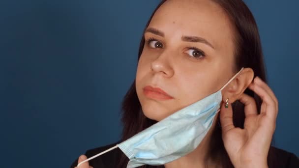 Retrato de una joven que se pone una máscara médica en la cara, estando aislada en casa sobre fondo azul. Mujer adulta protegiéndose de enfermedades. Concepto de amenaza de epidemia de coronavirus — Vídeo de stock