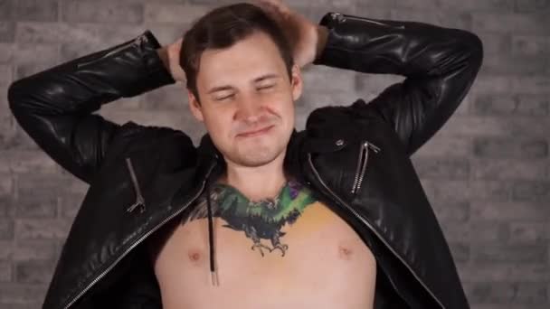 Красивый мужчина с татуировкой на груди в черной куртке позирует против серой кирпичной стены — стоковое видео