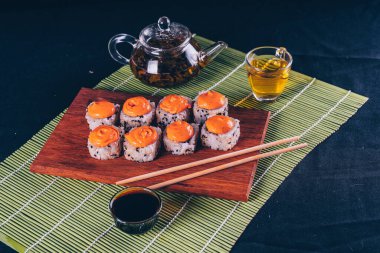 Ahşap tahtada baharatlı sos, çaydanlıkta yeşil çay ve dekoratif bambu peçetede fincan ile nefis dürümler. Siyah arka planda çubuklarla lezzetli suşi rulolarını kapatın..