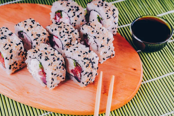 Крупный план вкусных суши-роллов на деревянной доске с соевым соусом и палочками на декоративной бамбуковой салфетке . — стоковое фото