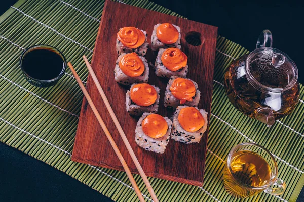 Deliziosi rotoli su tavola di legno con bacchette e tè verde in teiera su tovagliolo di bambù decorativo. Primo piano di gustosi rotoli di sushi sul tagliere su sfondo nero . — Foto Stock