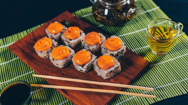 Deliciosos rollos sobre tabla de madera con palillos y té verde en tetera sobre servilleta de bambú decorativa. Primer plano de sabrosos rollos de sushi en la tabla de cortar sobre fondo negro . —  Fotos de Stock