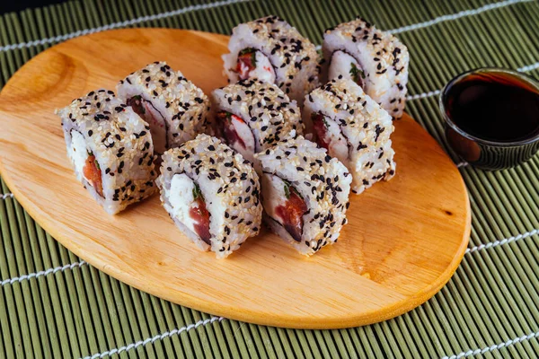 Крупный план вкусных суши-роллов на деревянной доске с соевым соусом и палочками на декоративной бамбуковой салфетке . — стоковое фото