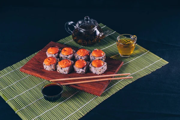 Deliziosi rotoli su tavola di legno con bacchette e tè verde in teiera su tovagliolo di bambù decorativo. Primo piano di gustosi rotoli di sushi sul tagliere su sfondo nero . — Foto Stock
