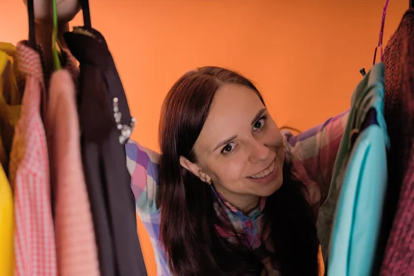 Młoda kobieta grymas stojąc między ubraniami w szafie. Dorosła kobieta wybiera rzeczy w sklepie. — Zdjęcie stockowe