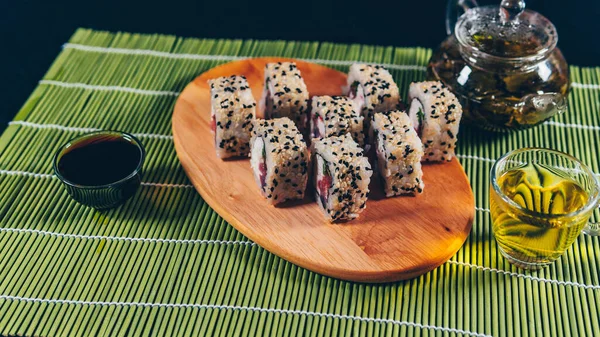 Deliciosos rollos sobre tabla de madera con salsa de soja y té verde sobre servilleta de bambú decorativa. Primer plano de sabrosos rollos de sushi con palillos en la tabla de cortar sobre fondo negro —  Fotos de Stock